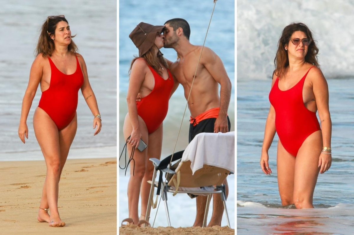 Fernanda Paes Leme de maiô vermelho e o namorado de bermuda preta com detalhe laranja, sem camisa, na areia da praia
