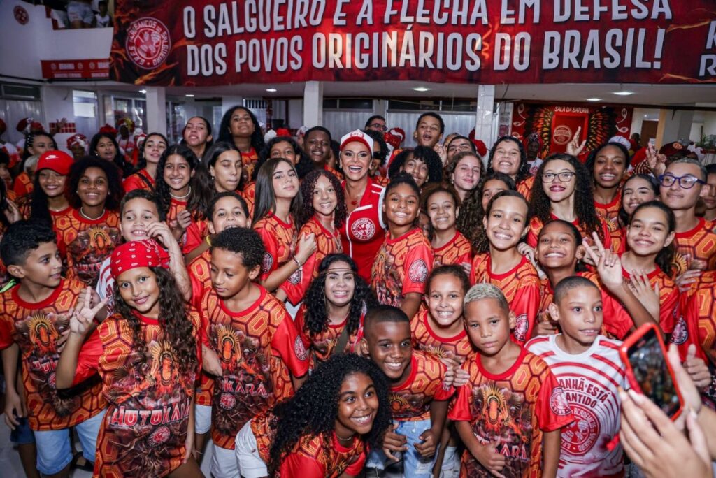 Viviane Araújo de macaquinho vermelho e branco, boné vermelho e branco, bota branca rodeada por crianças 