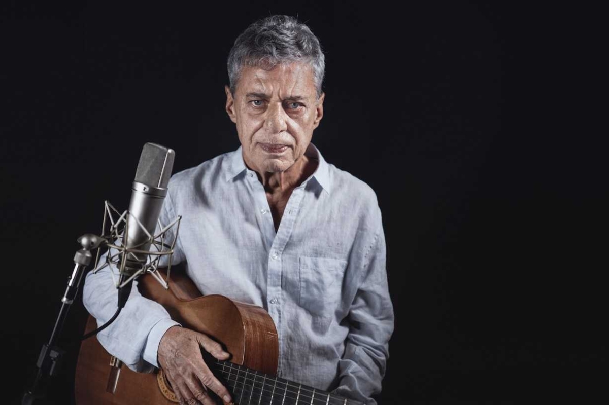 Chico Buarque diante de um microfone, segurando violão, de camisa branca de manga