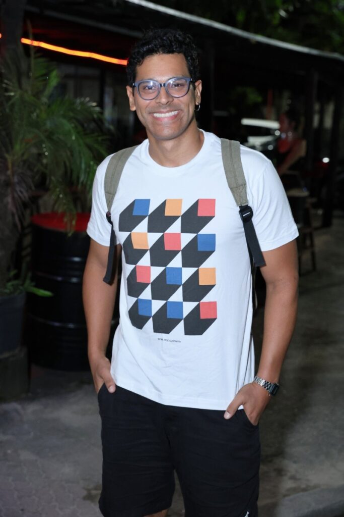 Renan Monteiro de camisa branca com estampa de quadrados coloridos, calça preta e mochila cinza 