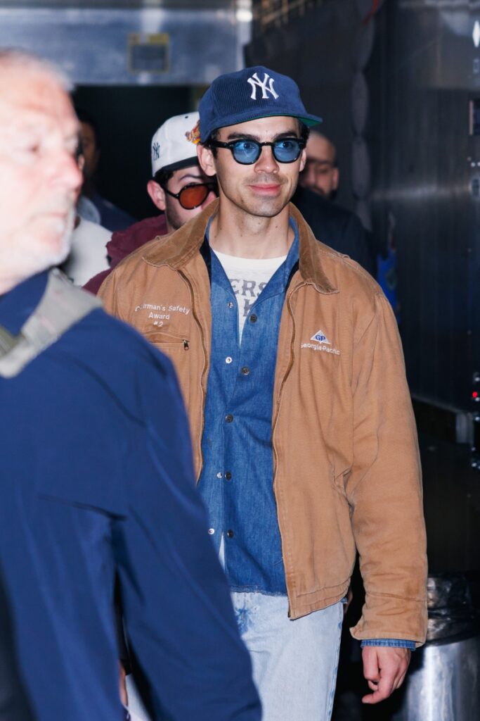 Joe Jonas de jaqueta marrom e camisa jeans, boné azul e óculos escuros 