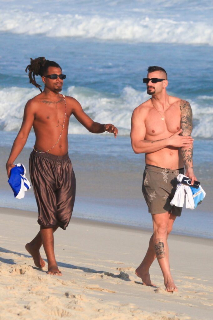 Luigi e Nizam na praia, ambos de bermuda e óculos escuros, andando na areia