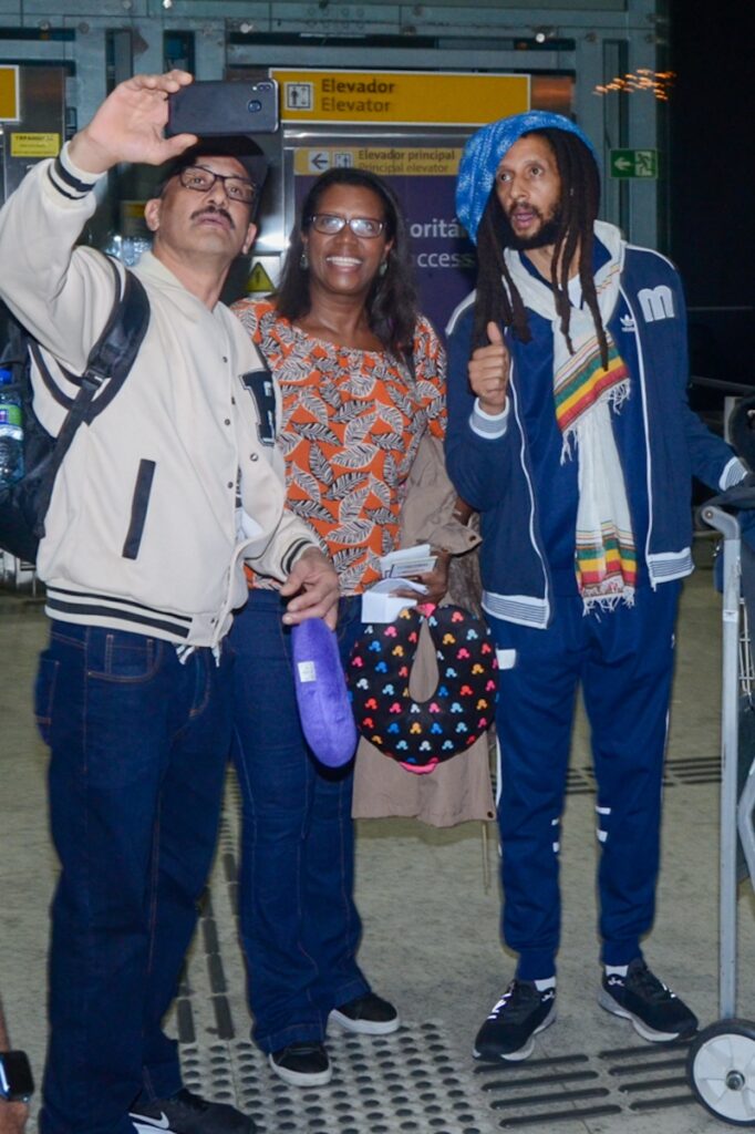 Julian Marley de casaco e calça azul, boina azul, no aeroporto  no aeroporto fazendo selfie com fãs 