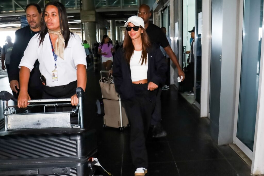 Anitta andado no aeroporto de conjunto de moletom preto, boné branco, camisa branca e óculos escuros 