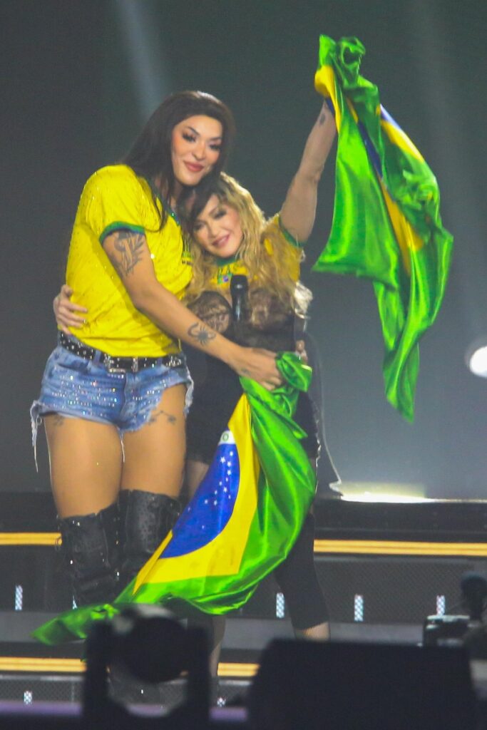 Madonna e Pabllo Vittar abraçadas, cada uma segurando com a bandeira do Brasil